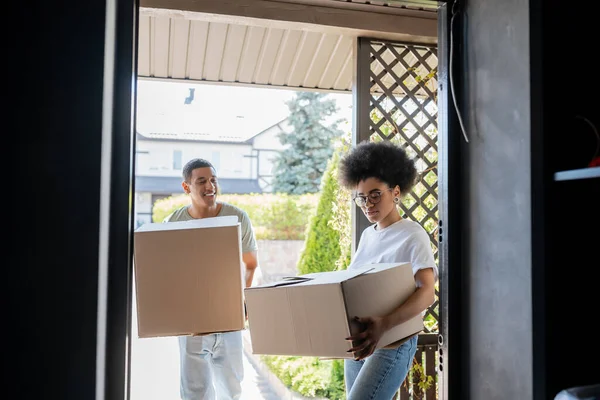Hombre afroamericano feliz sosteniendo la caja de cartón cerca de novia con el paquete y la puerta de la nueva casa - foto de stock