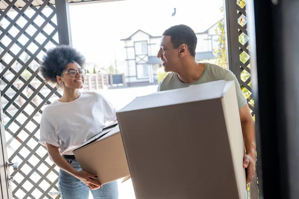 Pareja afroamericana positiva sosteniendo cajas de cartón cerca de la puerta de la nueva casa durante el movimiento - foto de stock