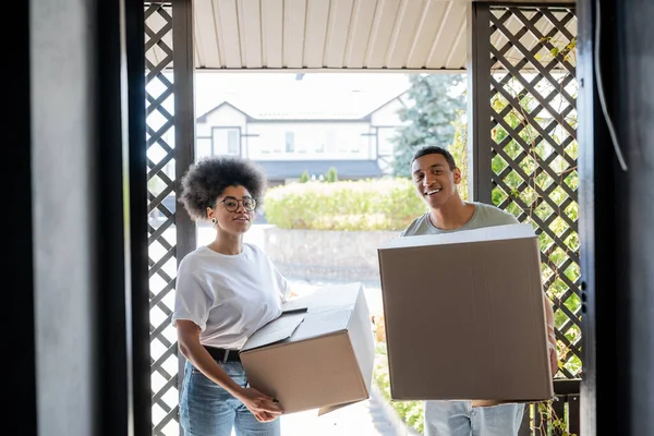Joyeux couple afro-américain avec des paquets regardant la caméra près de la porte de la nouvelle maison — Photo de stock
