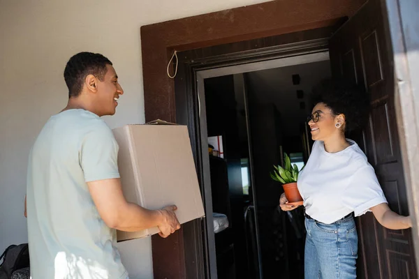 Allegra coppia afro-americana che tiene pianta e scatola di cartone vicino alla porta della nuova casa — Foto stock