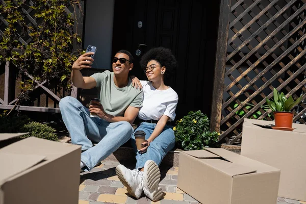 Улыбающаяся африканская американская пара делает селфи и держит кофе, чтобы подойти к коробкам и новому дому — стоковое фото