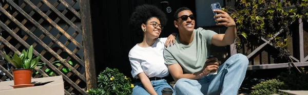 Couple afro-américain positif avec prise de café selfie près de boîtes en carton et près de la maison, bannière — Photo de stock