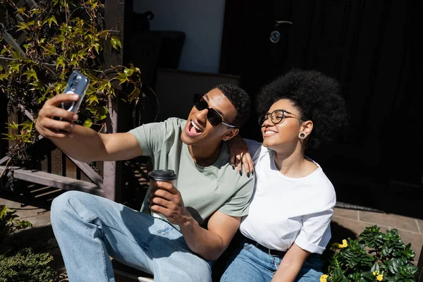 Весела афро-американська пара з кавою бере селфі на смартфон біля нового будинку — стокове фото