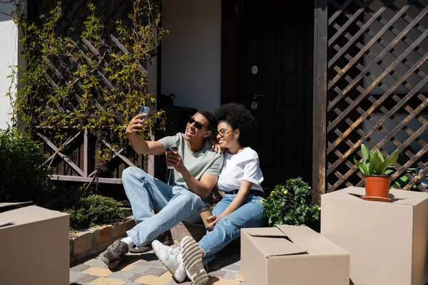 Sonriente afroamericano pareja con café para ir tomando selfie cerca de cajas y nueva casa - foto de stock