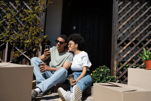 Улыбающаяся африканская американская пара с кофе, чтобы пойти с помощью смартфона рядом с коробками и новый дом — стоковое фото