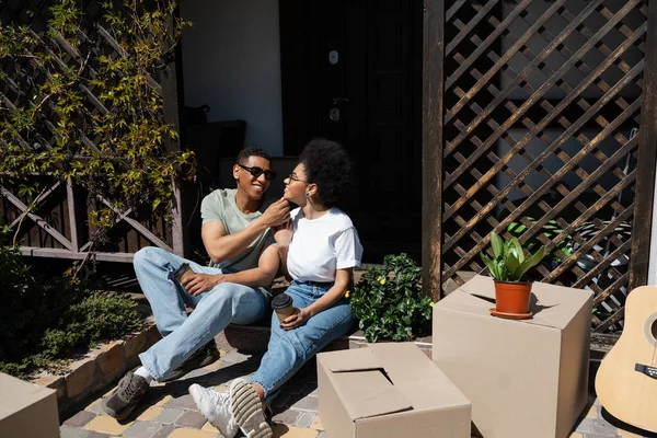Positif couple afro-américain avec du café pour aller assis près de boîtes en carton et nouvelle maison — Photo de stock