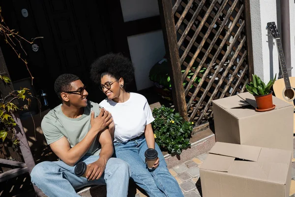 Alegre pareja afroamericana con café cogido de la mano cerca de cajas de cartón y nueva casa - foto de stock