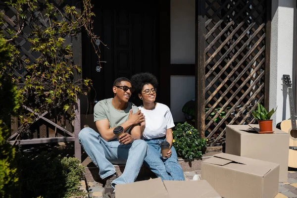 Sonriente pareja afroamericana con café hablando cerca de cajas de cartón en el porche de la nueva casa - foto de stock