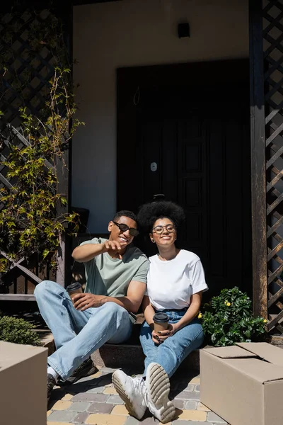 Souriant homme afro-américain avec café pointant vers des boîtes près de petite amie sur le porche de la nouvelle maison — Photo de stock
