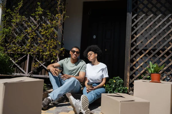 Щаслива афро-американська пара тримає каву і сидить біля пакетів і нового будинку — стокове фото