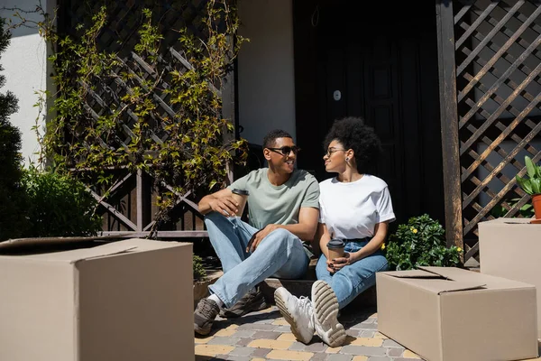 Улыбающийся африканский американец в солнцезащитных очках держит кофе и разговаривает с подругой возле нового дома — стоковое фото