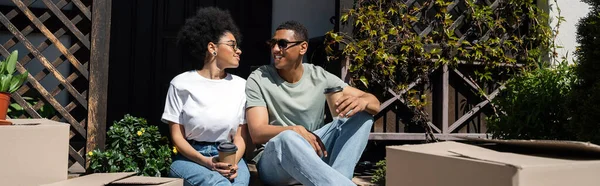 Heureux homme afro-américain parlant à sa petite amie avec du café près de boîtes en carton et nouvelle maison, bannière — Photo de stock