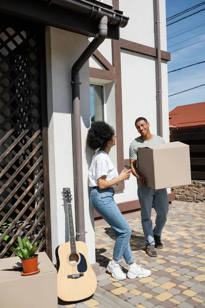 Smiling african american man holding cardboard box near girlfriend with coffee and new house — Stock Photo