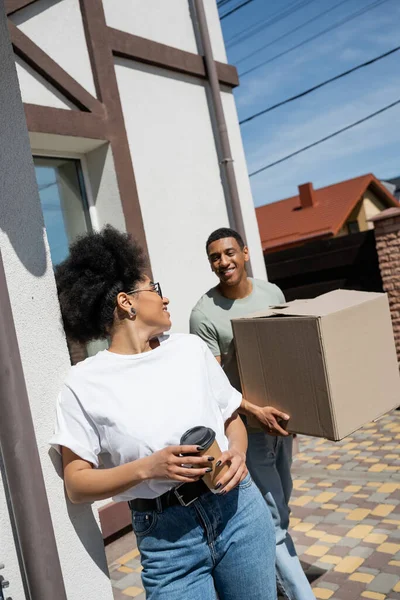 Sonriente afroamericana mujer sosteniendo café para ir cerca de novio con paquete y nueva casa - foto de stock