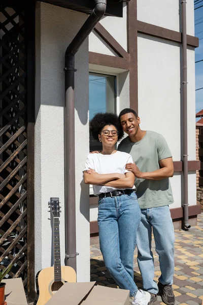 Sourire homme afro-américain étreignant petite amie près de la guitare acoustique et des boîtes près de la nouvelle maison — Photo de stock
