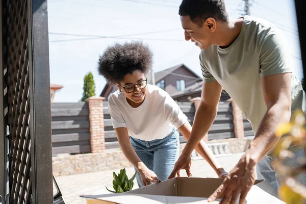 Cheerful african american woman unpacking carton box with boyfriend near new house outdoors — Stock Photo