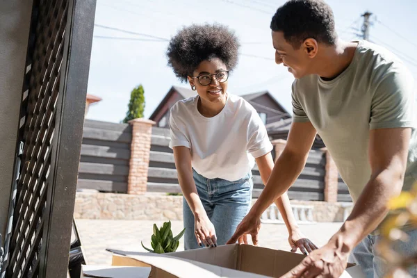 Positive african american couple talking while unpacking cardboard box near new house outdoors — Stock Photo