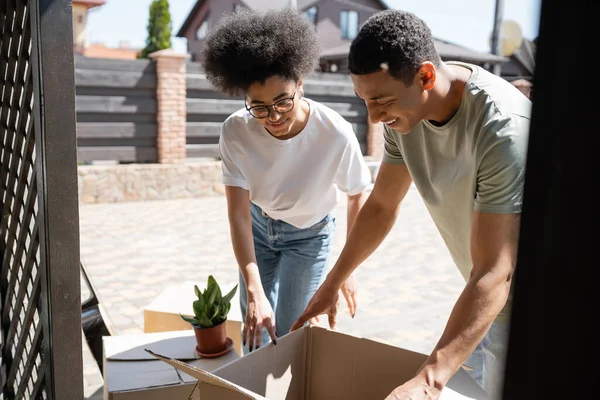 Souriant couple afro-américain déballer des boîtes en carton près des plantes et de la nouvelle maison — Photo de stock
