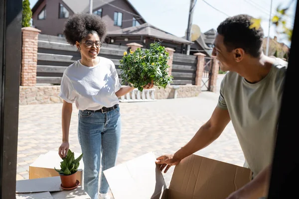 Femme afro-américaine souriante tenant une plante d'intérieur près de son petit ami et boîtes en carton pendant la réinstallation — Photo de stock