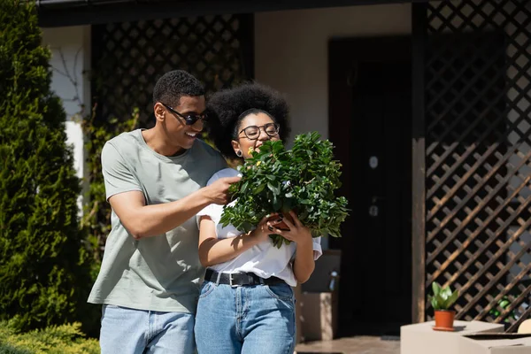 Cheerful african american couple holding houseplant near cardboard boxes and new house outdoors — Stock Photo