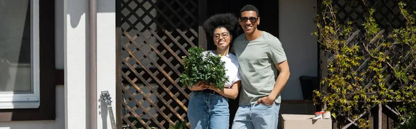 Coppia afro-americana positiva che tiene pianta d'appartamento vicino alla scatola di cartone e alla nuova casa, banner — Foto stock