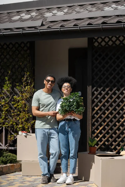 Щаслива афро-американська пара тримає кімнатну рослину біля пакетів і новий будинок на відкритому повітрі — стокове фото