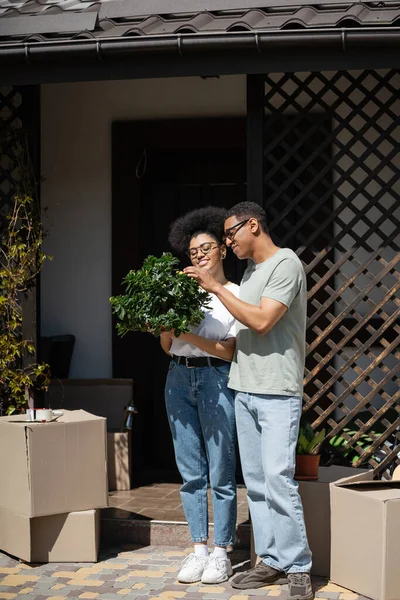 Alegre casal afro-americano olhando para houseplant perto de pacotes de papelão e nova casa — Fotografia de Stock