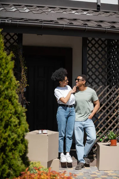 Улыбающаяся африканская американка трогает парня, стоя рядом с пакетами и новым домом — стоковое фото