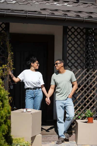 Positif couple afro-américain tenant la main près de boîtes en carton et nouvelle maison à l'extérieur — Photo de stock