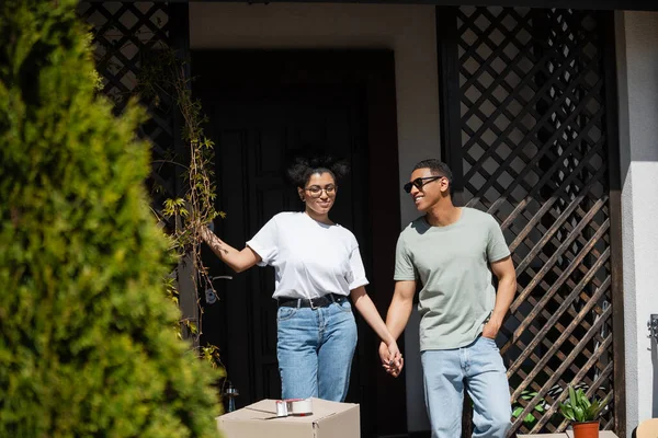 Улыбающаяся африканская американская пара держась за руки рядом с пакетом и новым домом на заднем плане — стоковое фото
