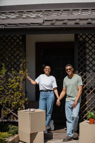 Felice coppia afro-americana che si tiene per mano vicino scatole di cartone e sul portico della nuova casa — Foto stock
