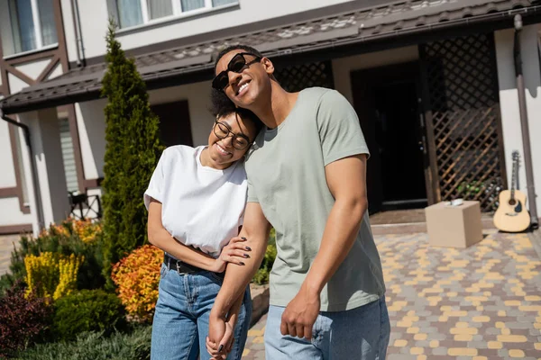 Freudiges afrikanisch-amerikanisches Paar hält Händchen, während es in der Nähe des neuen Hauses vor dem Hintergrund im Freien steht — Stockfoto