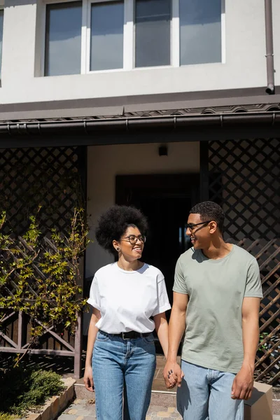 Lächelnder afrikanisch-amerikanischer Mann mit Sonnenbrille hält Hand seiner Freundin in der Nähe des neuen Hauses im Freien — Stockfoto