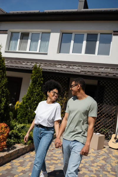Glückliches afrikanisch-amerikanisches Paar hält Händchen, während es in der Nähe des neuen Hauses im Hintergrund steht — Stockfoto