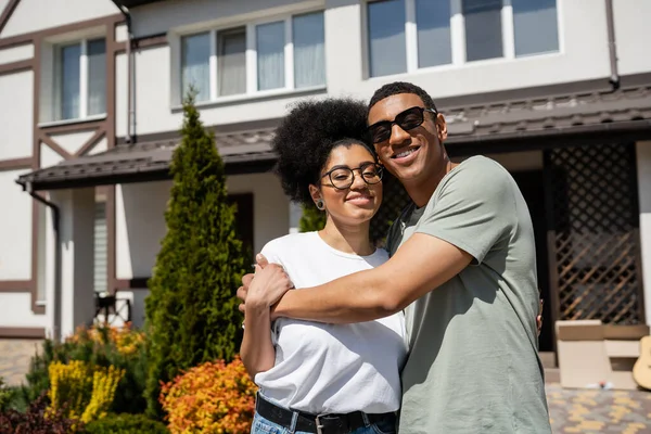 Sorridente uomo africano americano in occhiali da sole abbracciare fidanzata mentre in piedi vicino a nuova casa — Foto stock