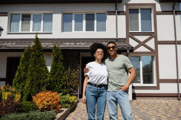 Positif couple afro-américain étreignant tout en se tenant près de la nouvelle maison sur fond — Photo de stock