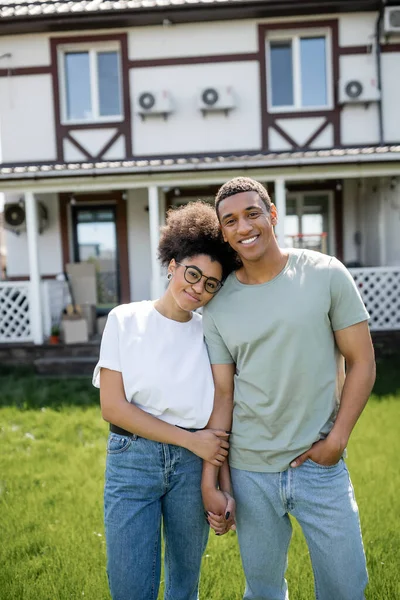 Улыбающаяся африканская американская пара, держащаяся за руки и смотрящая в камеру рядом с новым домом — стоковое фото