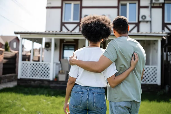 Rückansicht eines afrikanisch-amerikanischen Paares, das sich umarmt, während es in der Nähe eines neuen Hauses im Hintergrund steht — Stockfoto