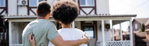 Vista trasera de la pareja afroamericana abrazándose mientras está de pie cerca de la nueva casa al aire libre, pancarta - foto de stock