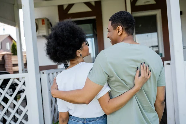 Sorridente casal afro-americano abraçando e olhando um para o outro perto de casa nova — Fotografia de Stock
