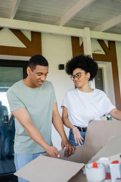 African american woman pointing at carton box near boyfriend during moving in new house — Stock Photo