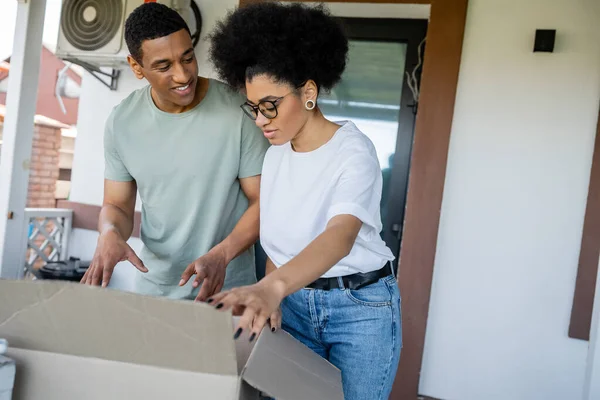 Femme afro-américaine déballage boîte en carton près petit ami souriant lors de la réinstallation dans une nouvelle maison — Photo de stock