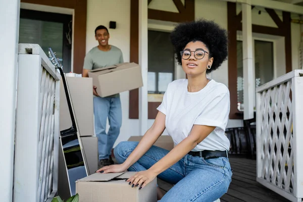 Mulher americana africana olhando para a câmera perto de caixas de papelão e namorado durante a mudança na nova casa — Fotografia de Stock