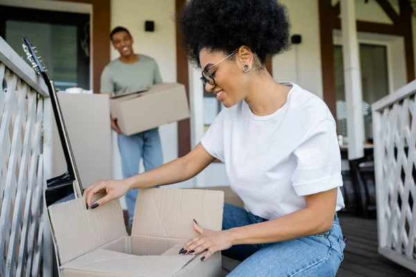 Весела афроамериканка розпаковує картонну коробку біля розмитого хлопця і нового будинку — стокове фото