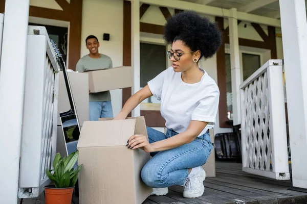Афроамериканська жінка розпаковує картонну коробку біля розмитого хлопця і нового будинку — стокове фото