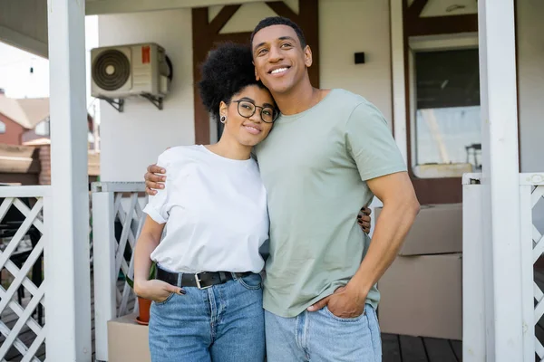 Positive african american couple hugging and posing near blurred new house on background — Stock Photo