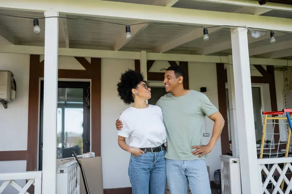 Happy african american couple embracing while standing on porch near new house outdoors — Stock Photo