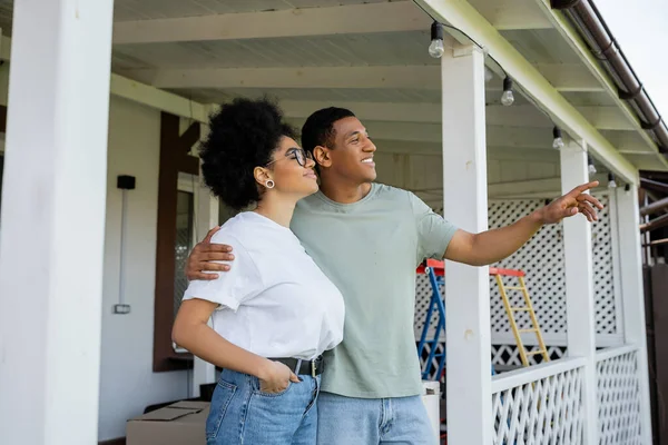 Souriant homme afro-américain étreignant et pointant près de petite amie sur le porche de la nouvelle maison — Photo de stock