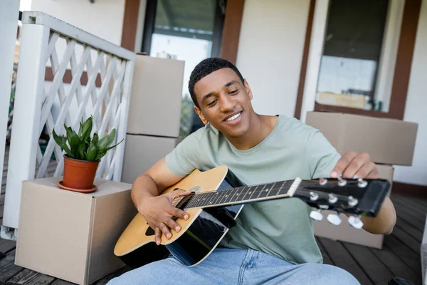 Улыбающийся африканский американец, играющий на акустической гитаре возле коробок и нового дома — стоковое фото