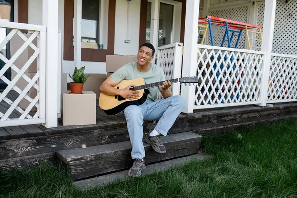 Веселый африканский американец, играющий на акустической гитаре на крыльце возле коробок и нового дома — стоковое фото